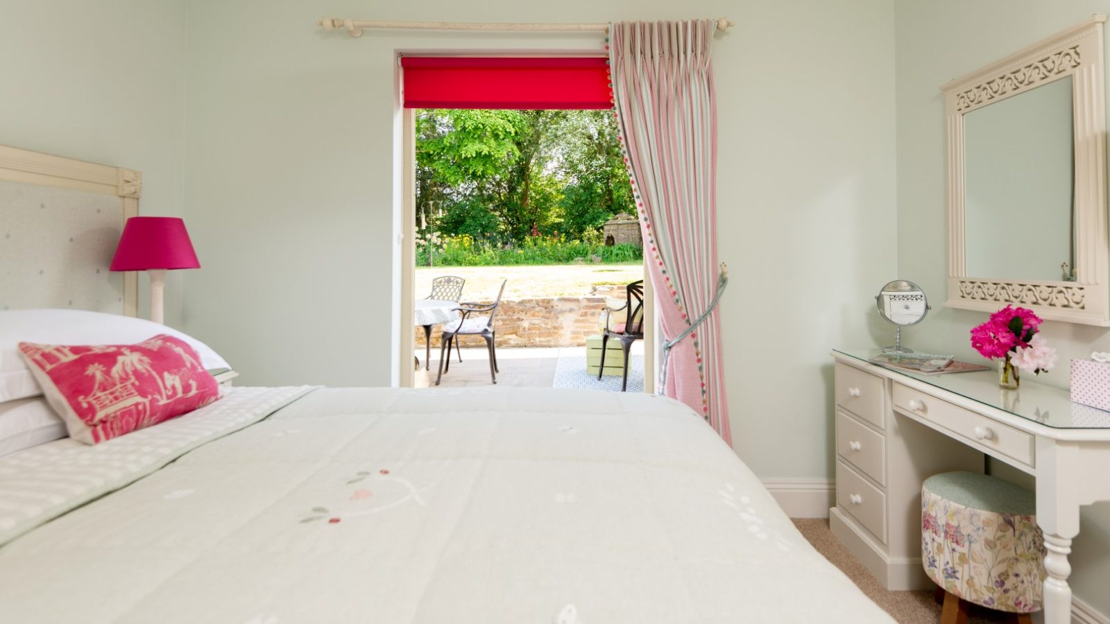 Sara's Cottage features a cozy bedroom with a pastel green color scheme, including a large bed with a pastel green quilt, a pink pillow, and a matching lamp. A vanity with a mirror and flowers sits to the right. The room also boasts a glass door leading to a patio with outdoor furniture.