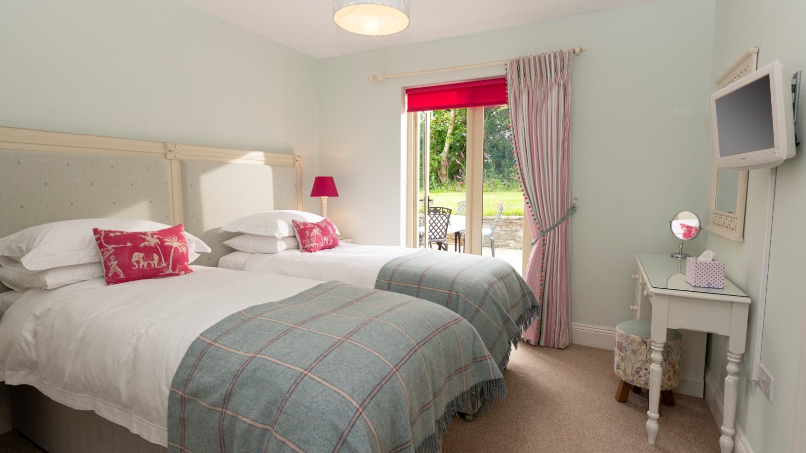 Sara's Cottage offers a cozy bedroom with two single beds covered in white and blue checkered bedding, a small desk with a mirror and chair, a wall-mounted TV, and a window adorned with pink curtains leading to an outdoor garden area. The room features soft, neutral wall colors.