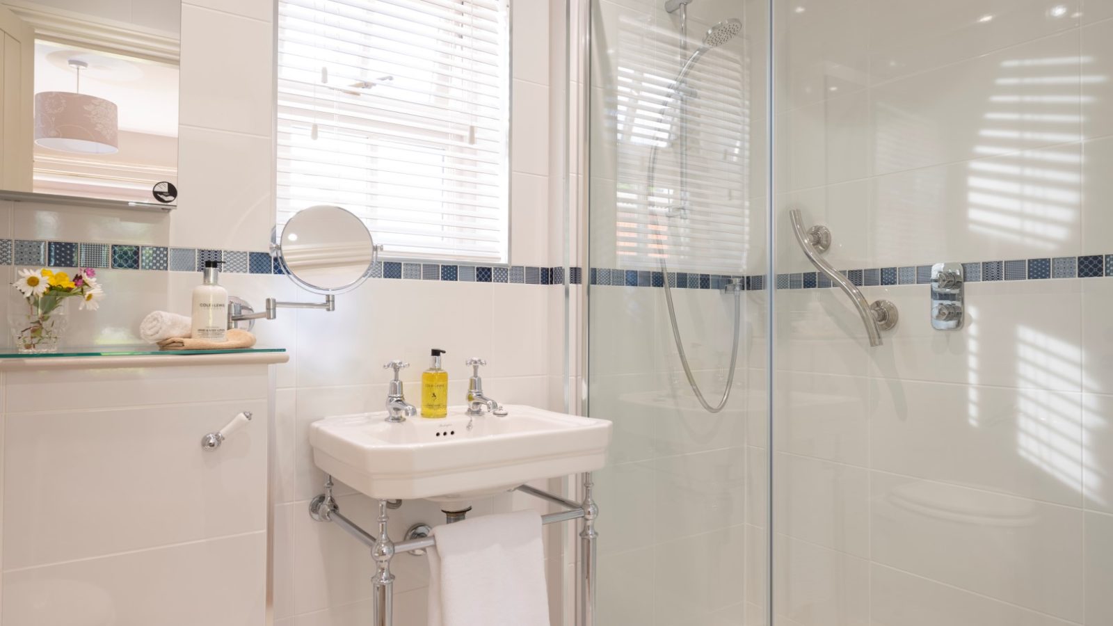 A modern bathroom in Sara's Cottage features a glass-enclosed shower with dual shower heads, a white ceramic sink with double faucets, a round mirror, and a window with white blinds. A small countertop next to the sink displays a soap dispenser, flowers, and neatly folded towels.