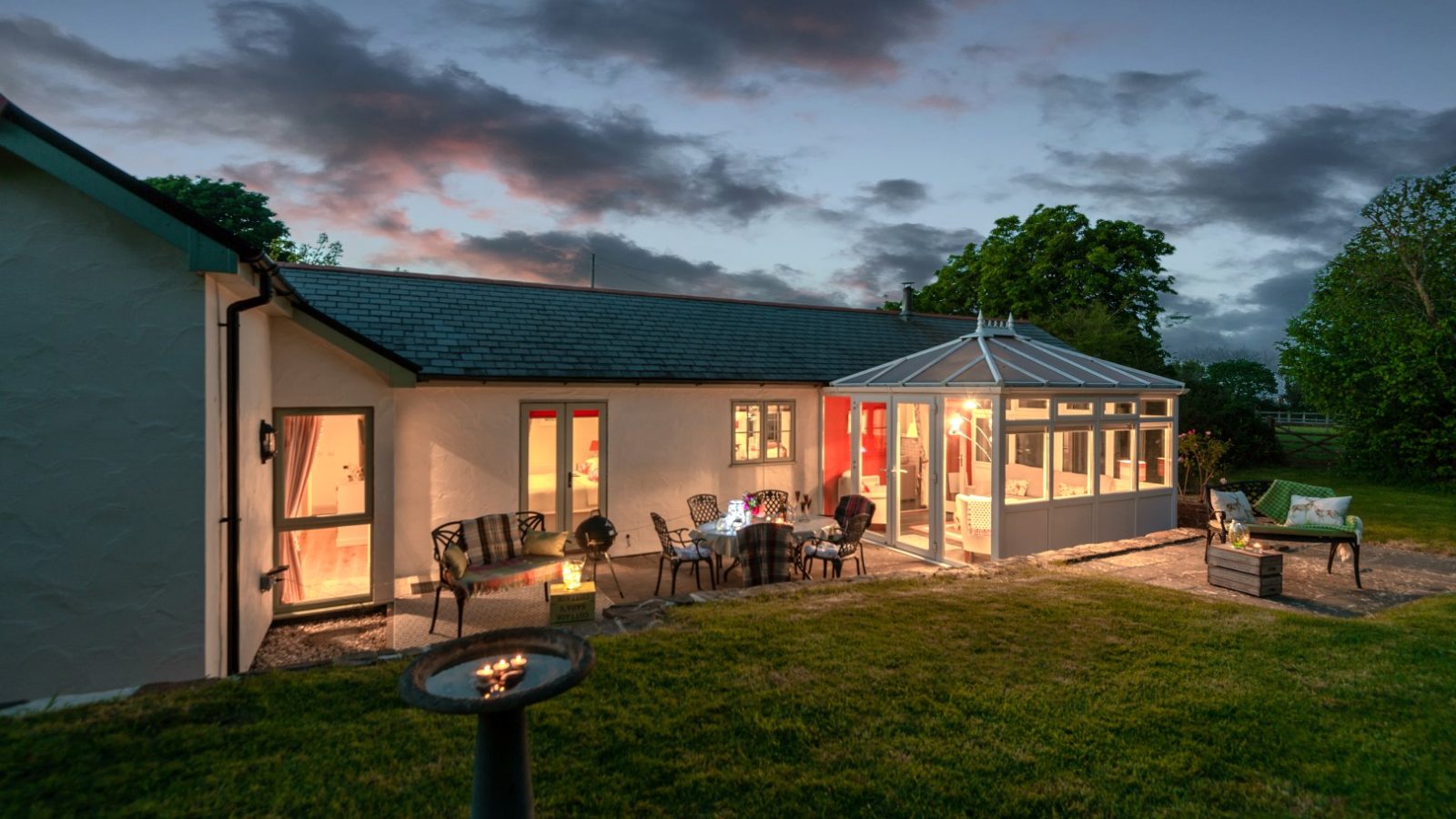 Sara's Cottage, a modern house with a glass conservatory, is illuminated at dusk. There is a patio area with outdoor furniture and a lit fire pit in the foreground. The surrounding lawn is neatly maintained, and a few trees are visible in the background.