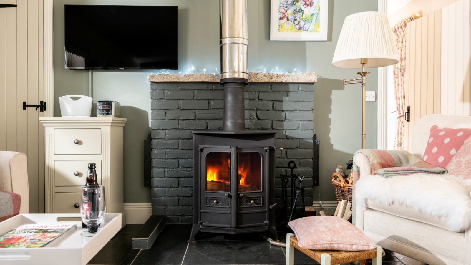 Sara's Cottage boasts a cozy living room featuring a burning fireplace with a black metal stove set against a painted brick wall. A TV and artwork hang above. The space includes comfy seating, a white side table with a bottle and magazine, a floor lamp, and soft cushions.