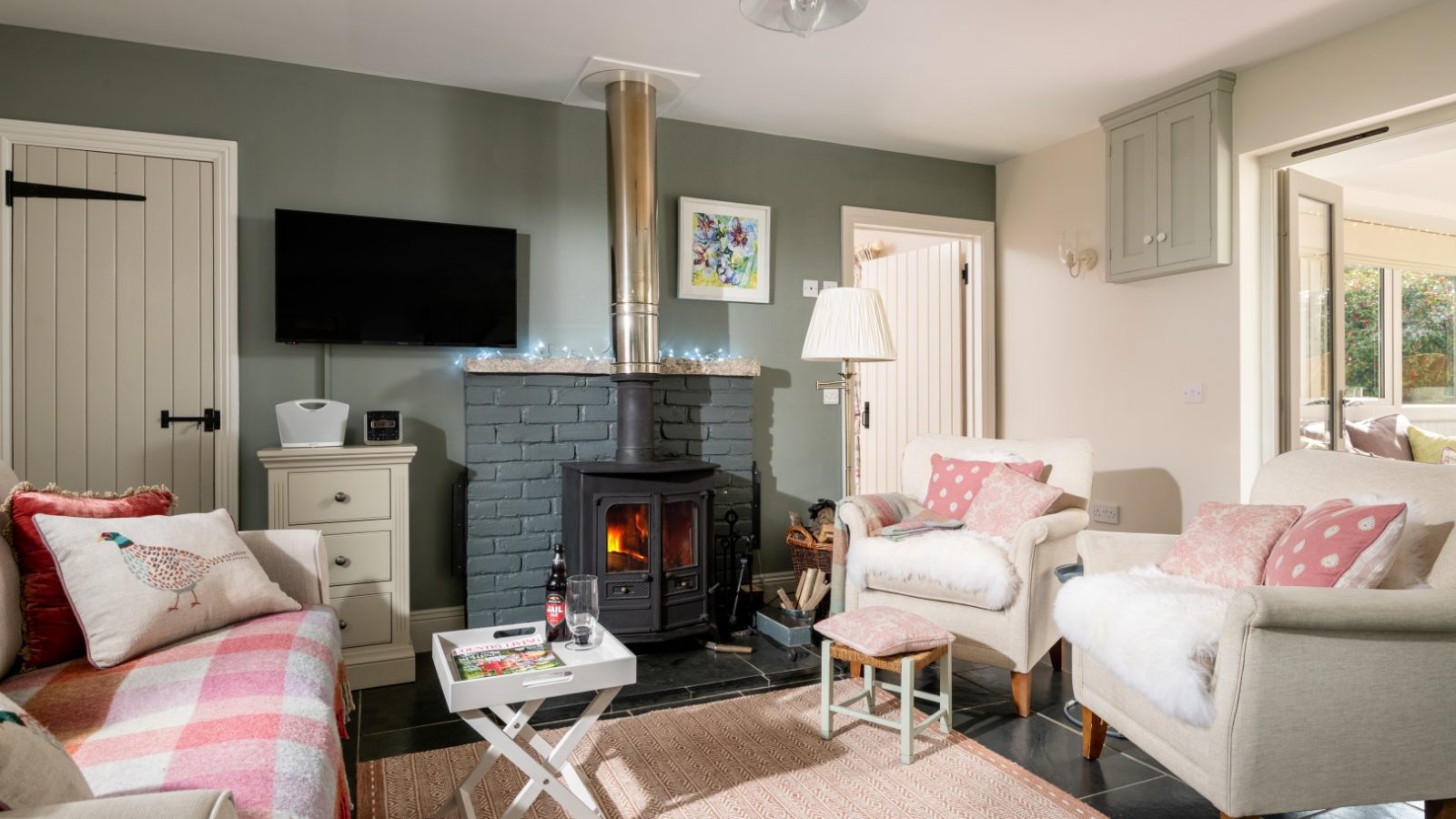 Sara's Cottage features a cozy living room with a wood-burning stove, two armchairs, and a sofa adorned with pink and white cushions. A small coffee table with magazines and a bottle sits on a patterned rug. A flat-screen TV is mounted on the wall, and a door leads to another room.