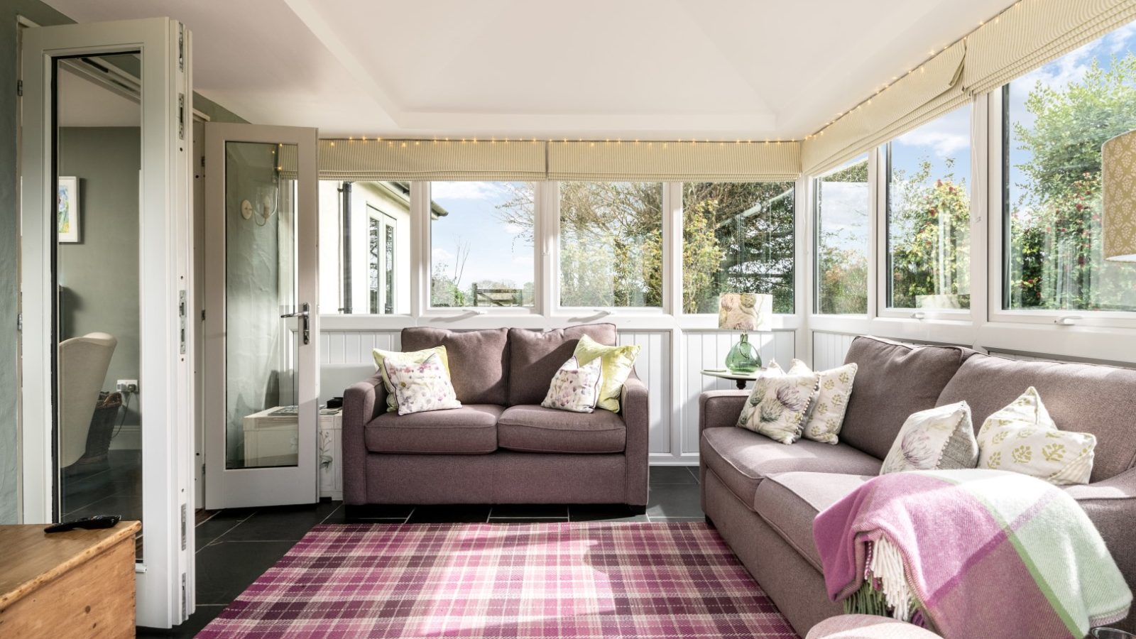 Sara's Cottage boasts a cozy sunroom with two grey couches adorned with patterned cushions. A pink and green plaid blanket is draped over one couch, while a pink and purple plaid rug covers the floor. Large windows bring in natural light, and a glass door opens to a view of greenery outside.