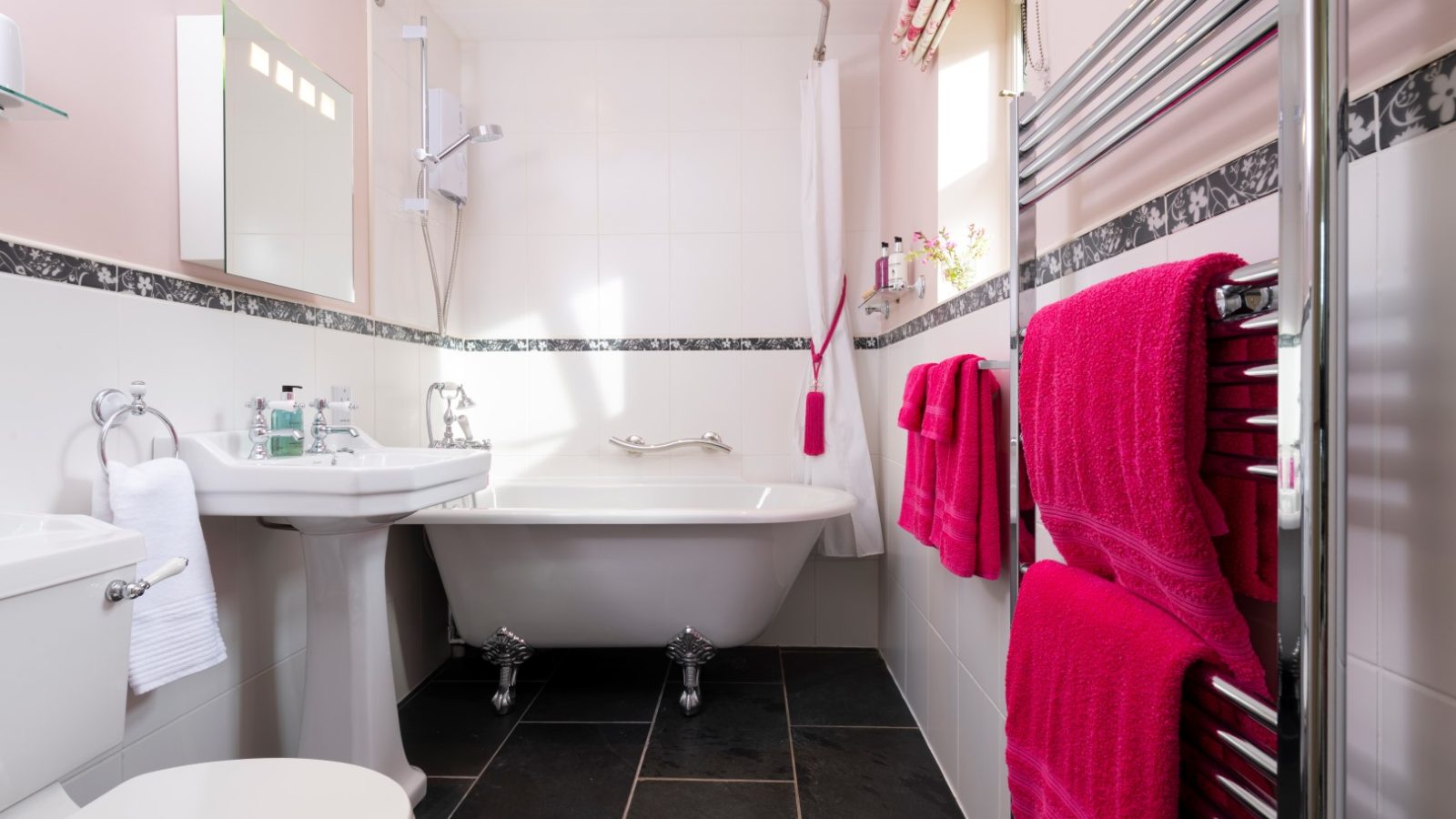 A modern bathroom in Sara's Cottage features a white bathtub, silver fixtures, and a showerhead. There's a white sink with toiletries on the counter, a toilet, and a heated towel rail with red towels. The walls are white with black tile accents, and the floor has dark tiles.