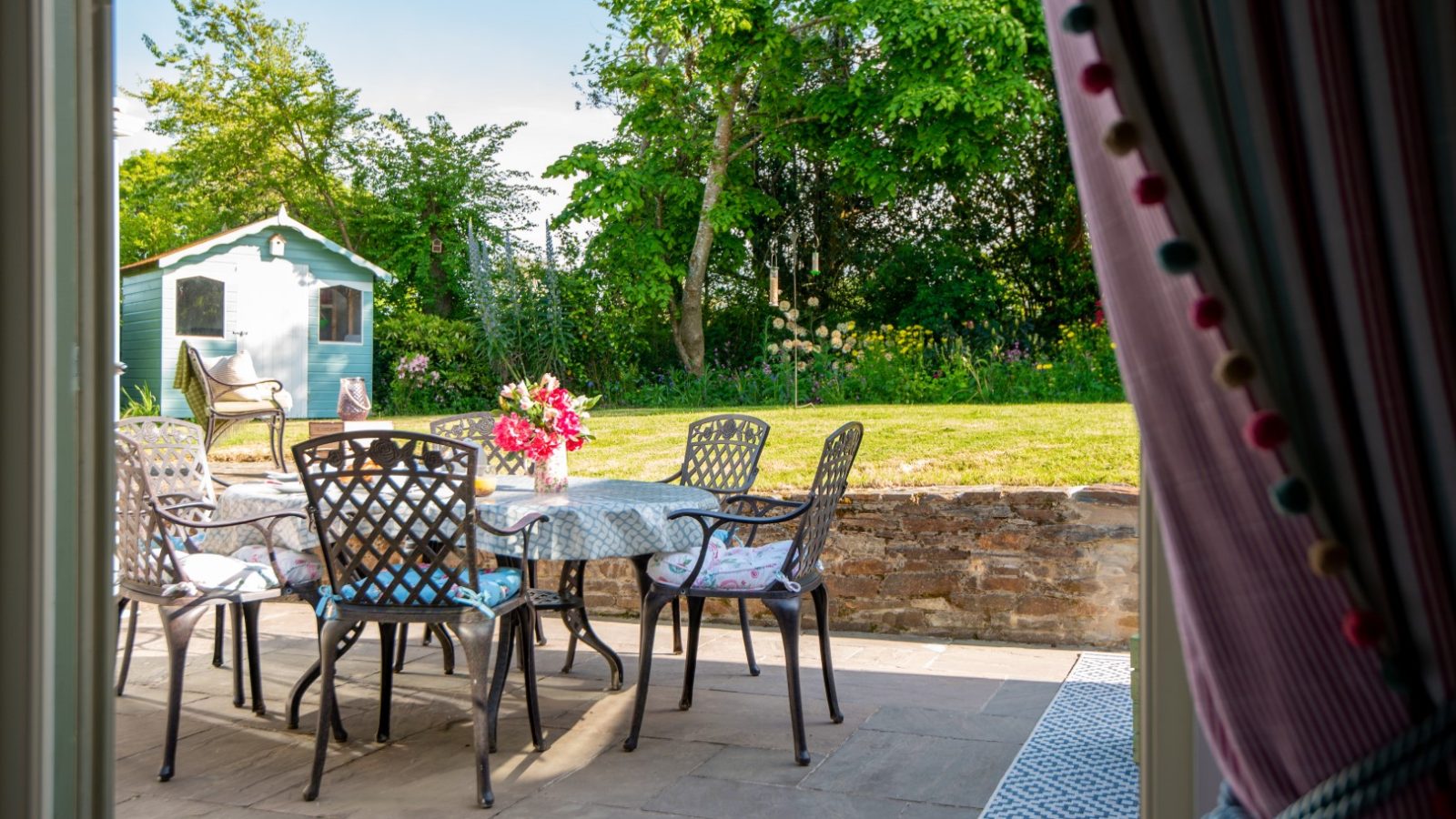 Sara's Cottage offers a cozy outdoor patio featuring a round glass table surrounded by six wrought-iron chairs, each with floral cushions. A small bouquet of flowers sits on the table. The patio overlooks a lush garden with a small blue shed, framed by colorful curtains with pom-poms.