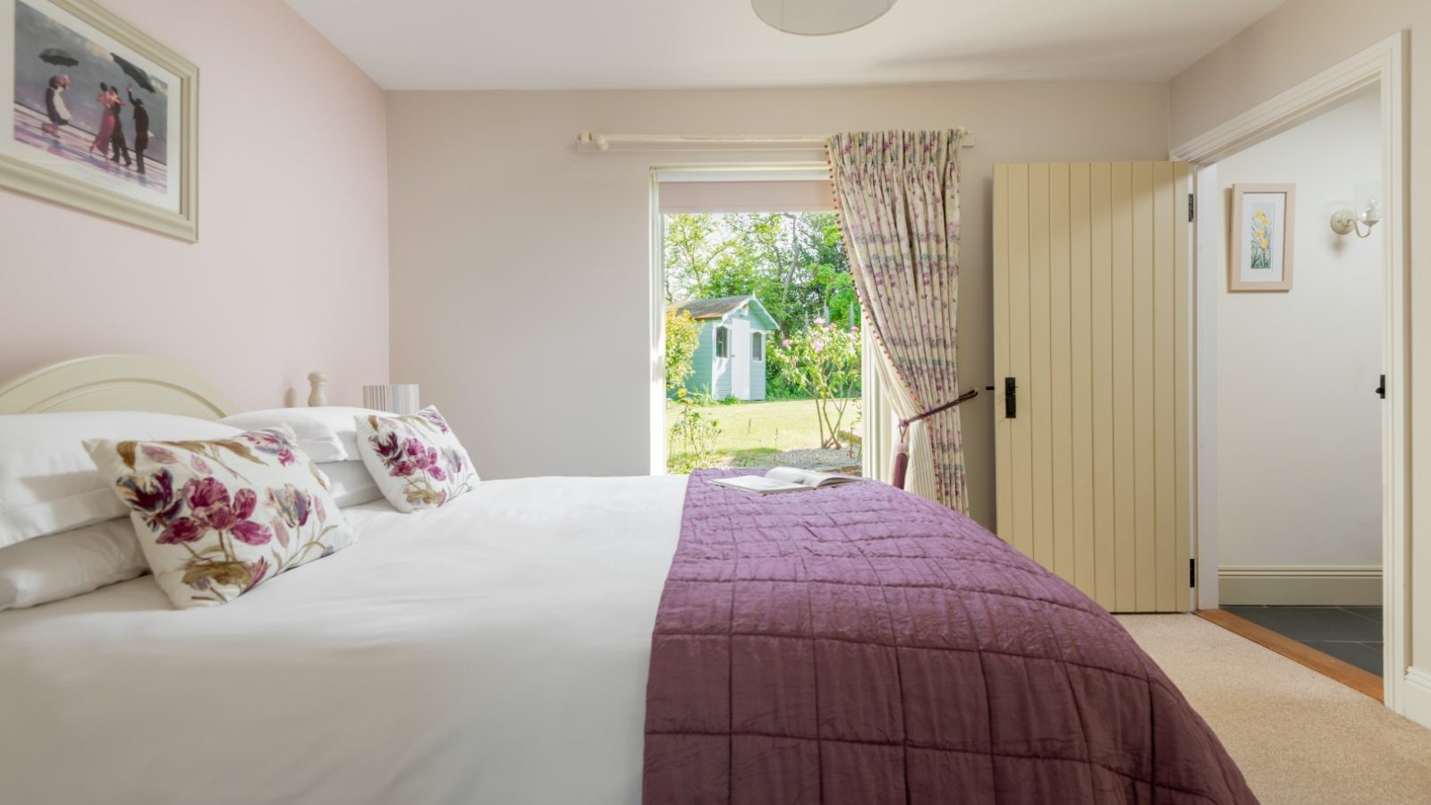 Sara's Cottage offers a cozy bedroom with a large bed, decorated with white and floral pillows and a purple quilt. The room has cream-colored walls and curtains. One door leads to a garden where a small green shed is visible, while another door connects to an adjoining room.