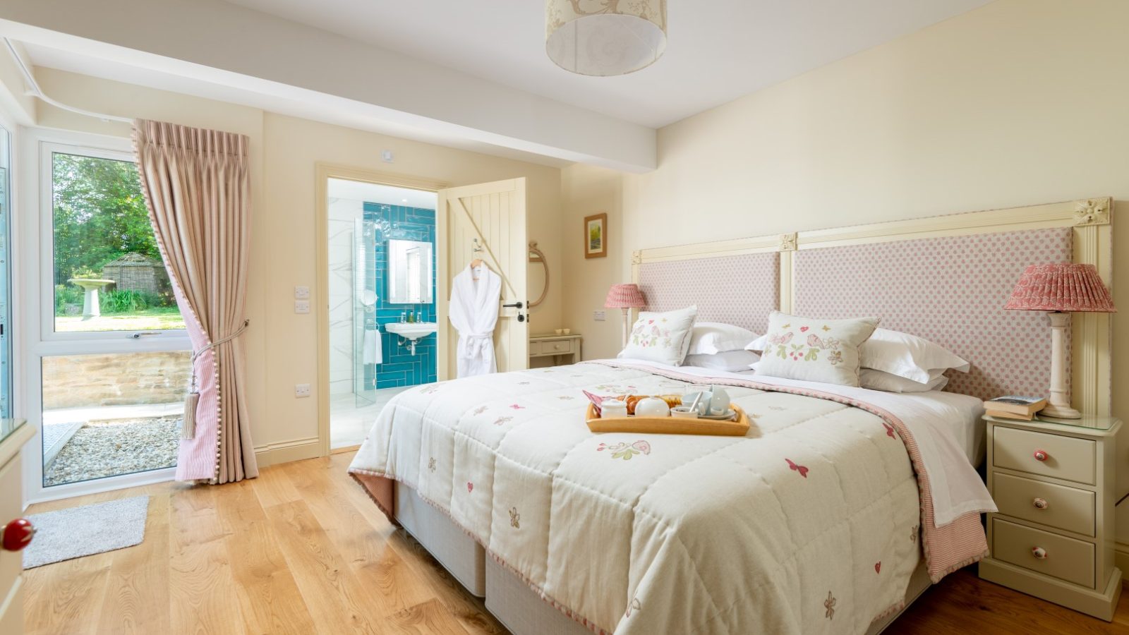 Sara's Cottage features a cozy, well-lit bedroom with a large double bed adorned with a beige and pink patterned quilt. Two bedside tables with matching lamps flank the bed. A wooden floor enhances the warmth of the room, which leads to a bathroom with an open door displaying a robe.