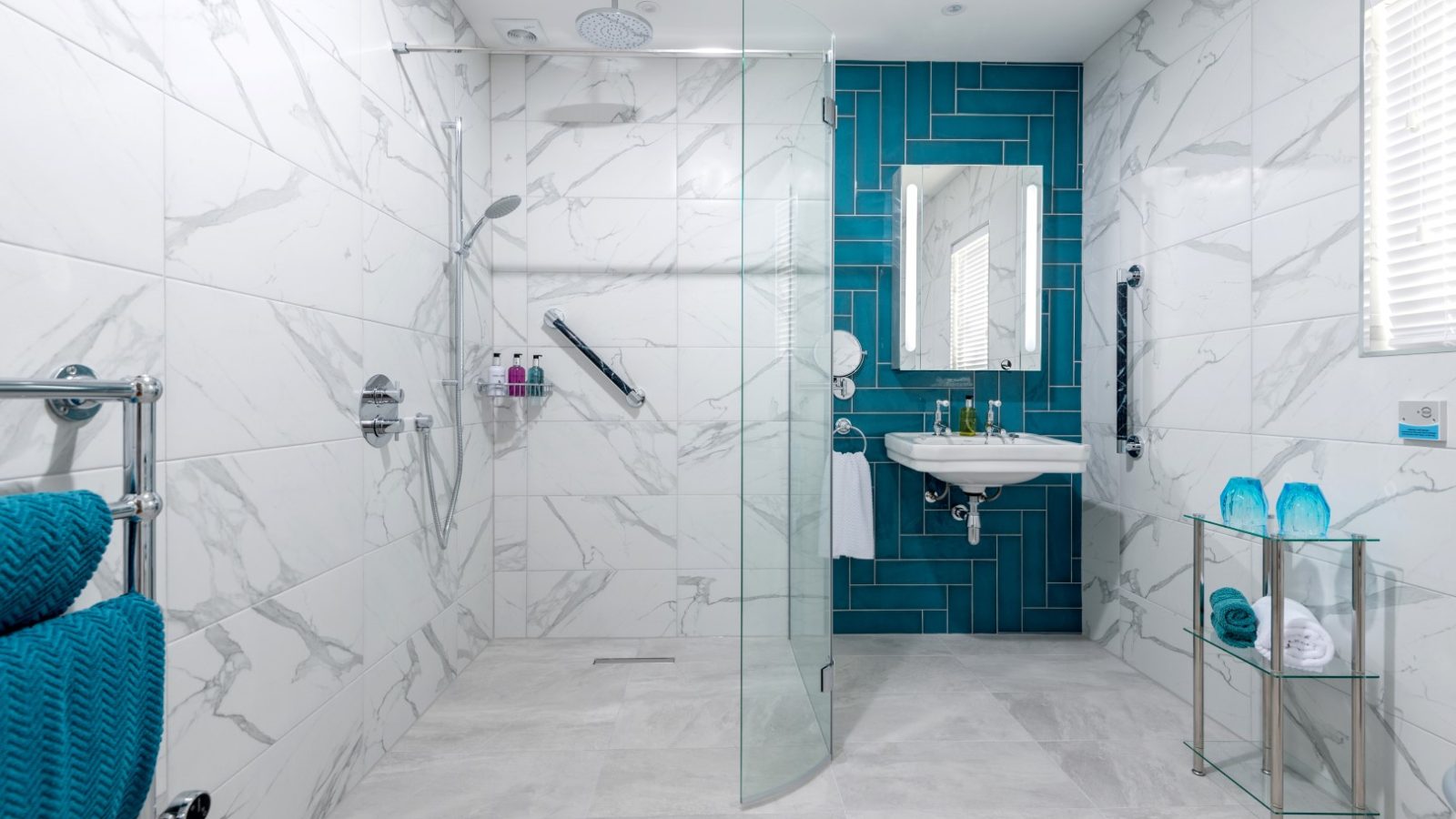 Sara's Cottage features a modern bathroom with white marble-patterned tiles, a glass shower enclosure, and a blue herringbone-tiled accent wall. The shower area boasts a rain showerhead, handheld shower, and grab bars. A wall-mounted sink sits beside a mirror, complemented by a heated towel rack with folded teal towels.