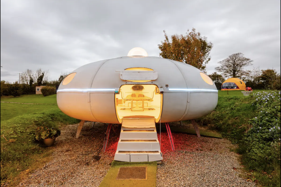 A modern, futuristic pod-like house with a circular shape and a glowing yellow interior. Elevated on stilts with stairs leading to the entrance, it looks like a UFO set in a grassy outdoor area. Another similar pod is visible in the background under an overcast sky—perfect for Apple Camping Adventures.