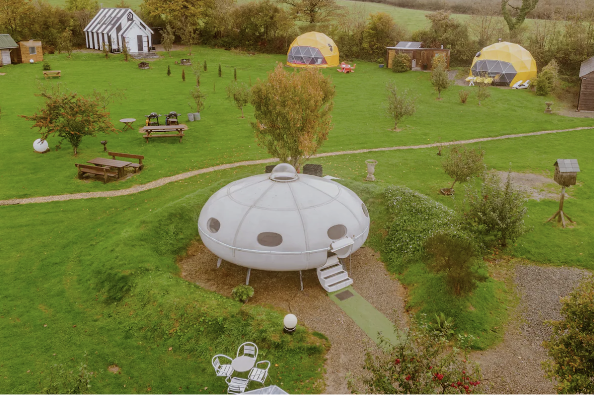 Embark on an Apple Camping Adventure at this scenic glamping site featuring unique accommodations, including a white, UFO-shaped pod with stairs, two yellow dome structures, and a striped hut. The site is surrounded by trees and green grass with pathways and picnic tables scattered throughout.