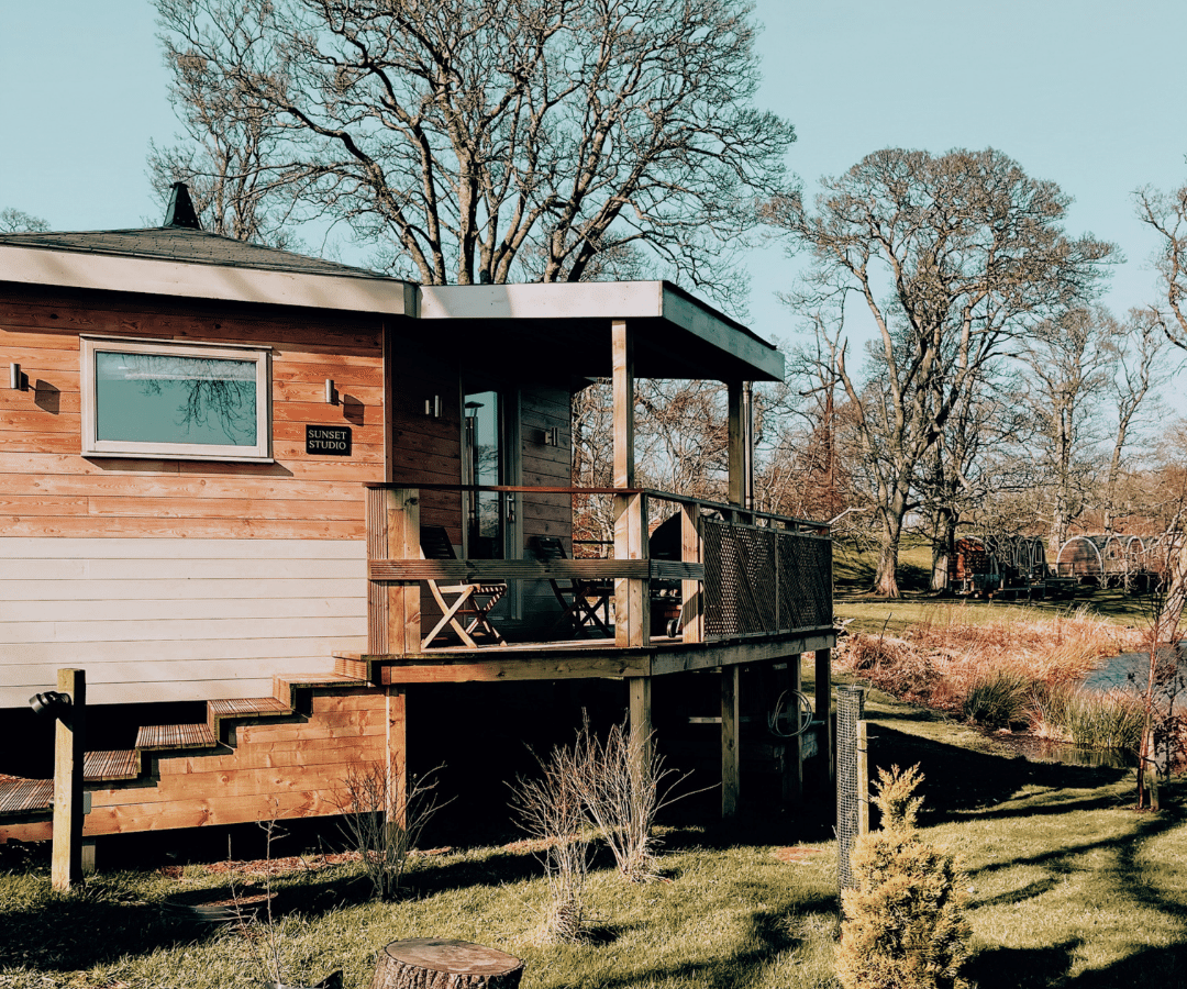 A cozy cabin with a wooden porch is nestled in the heart of Blossom Plantation, surrounded by a peaceful, natural landscape. Leafless trees and a small pond dot the background under a clear blue sky. The porch features a small table and chairs, while grass and shrubs blanket the ground.