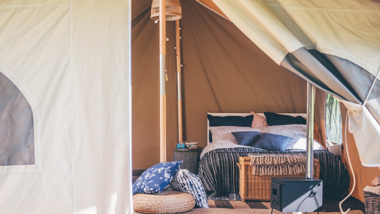 A cozy, well-furnished tent interior with a neatly made bed adorned with pillows and blankets. There is a small wooden nightstand, a woven basket, a pouf, and a tiny black stove, creating a warm and inviting atmosphere. Natural light filters through the tent in typical Nantseren fashion.