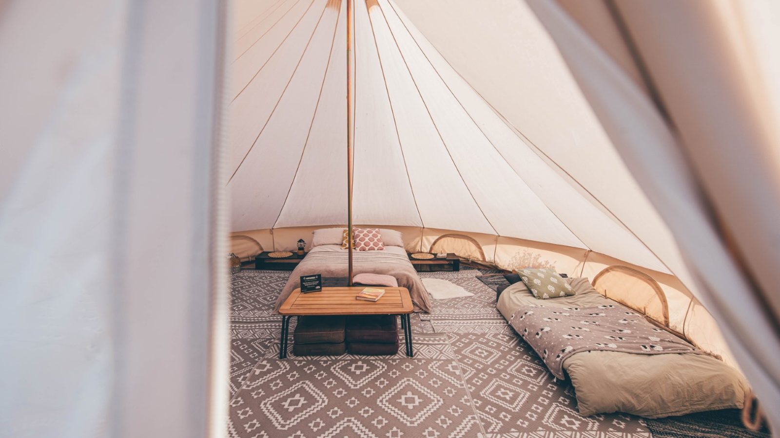 Interior of a Nantseren tent with a cozy setup, including patterned rugs, a central wooden table with books, a sofa with cushions, and a low bed with a blanket. The tent has a soft cream-colored canopy supported by a central pole, creating a warm, inviting atmosphere.