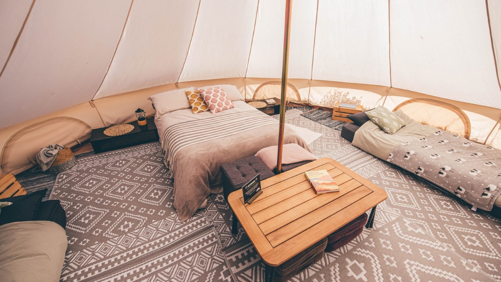 A cozy glamping tent at Nantseren with a large bed, a small bed, a wooden coffee table, and a patterned rug. The tent has soft, natural lighting and is decorated with cushions, blankets, and books, providing a comfortable and inviting atmosphere.