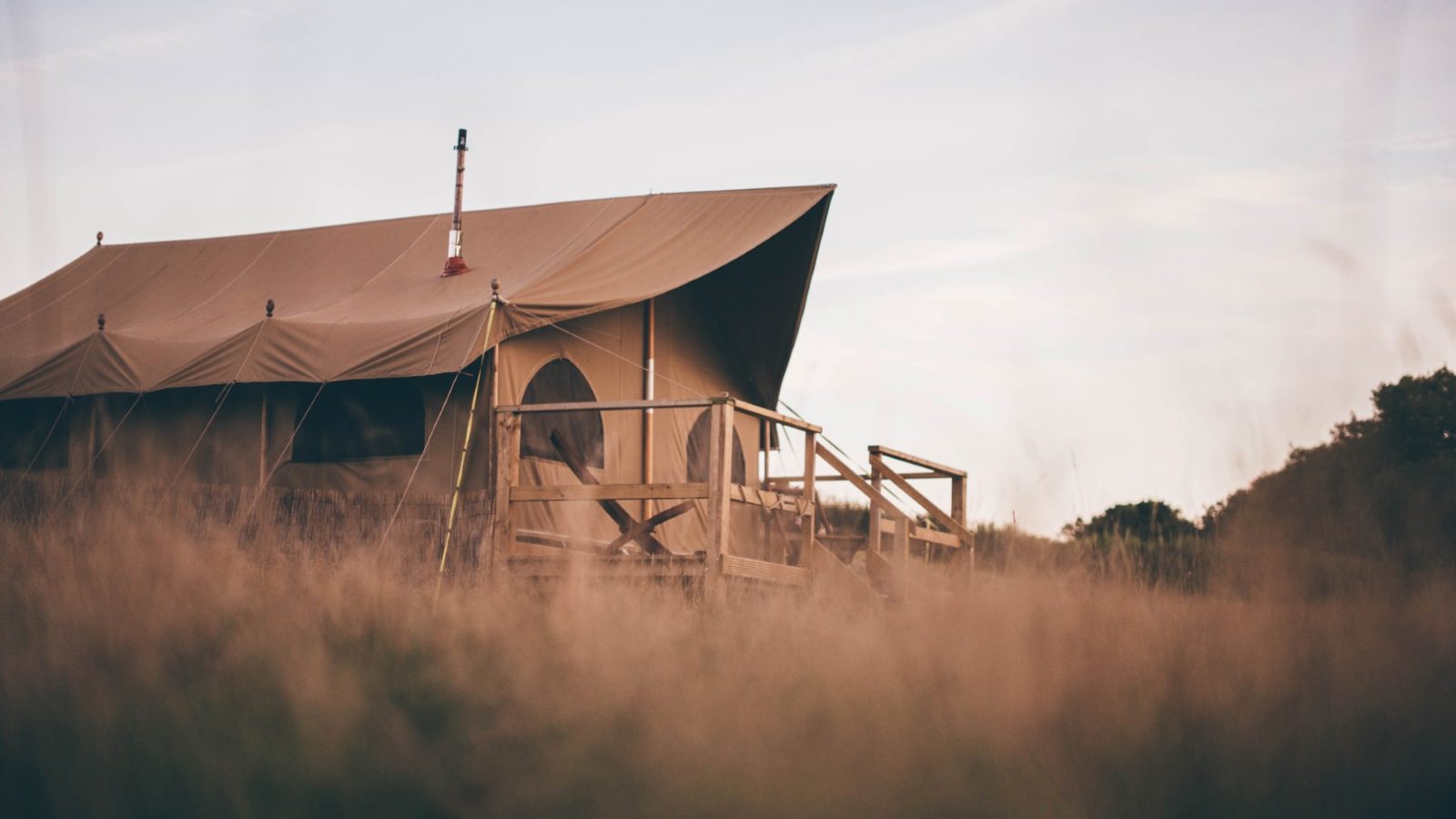 A large canvas tent is set up on a wooden platform in an open, grassy field. The tent, reminiscent of the ones used in Nantseren, features a chimney pipe, indicating it may have a wood stove inside. The scene is tranquil with soft, natural light, and some trees are visible in the background.