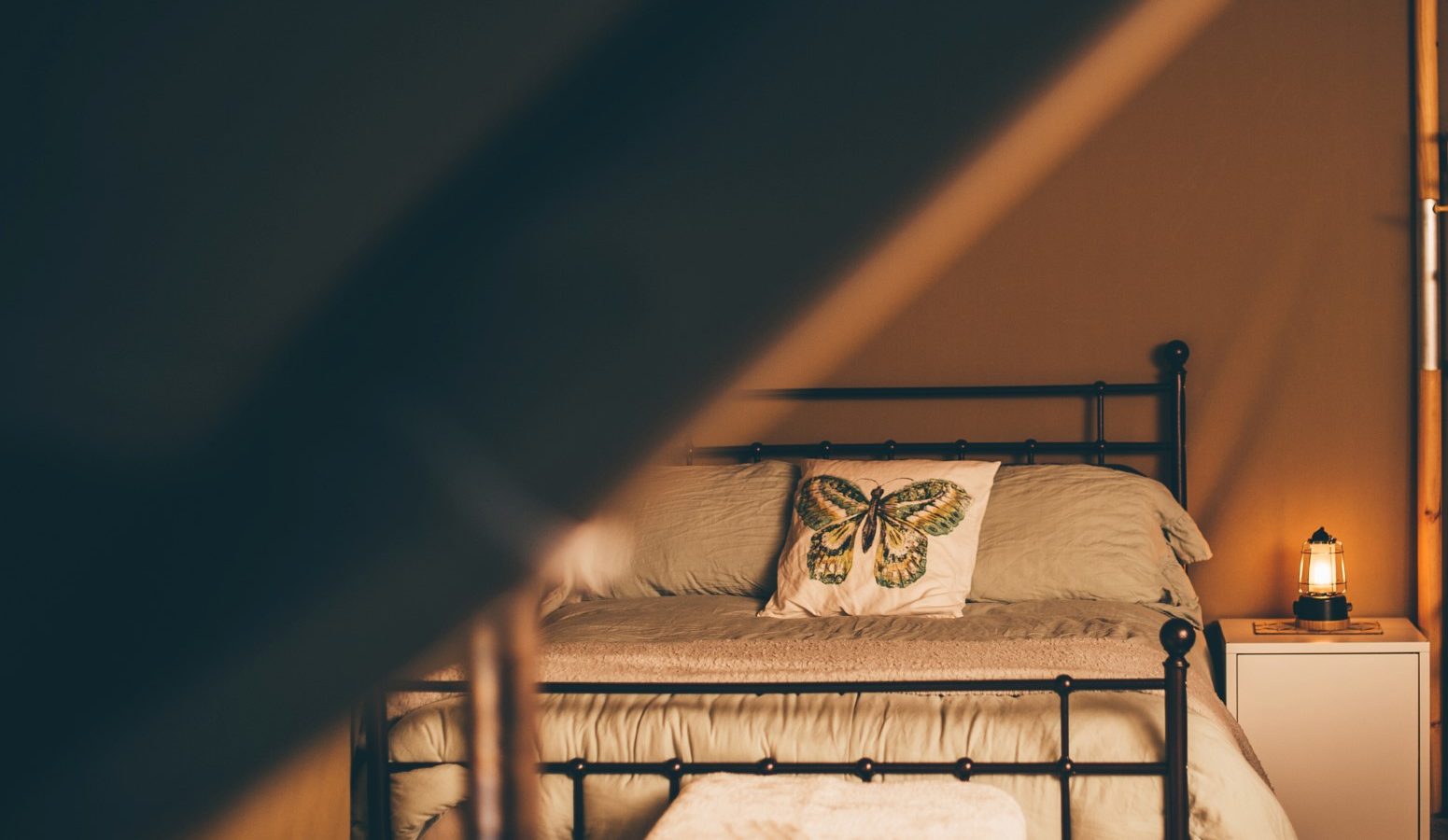 Cozy bedroom with a metal-framed bed adorned with pale green bedding and a butterfly-themed pillow. A small nightstand beside the bed features a Nantseren lantern-style lamp. The warm lighting and simple decor create a serene and inviting atmosphere.