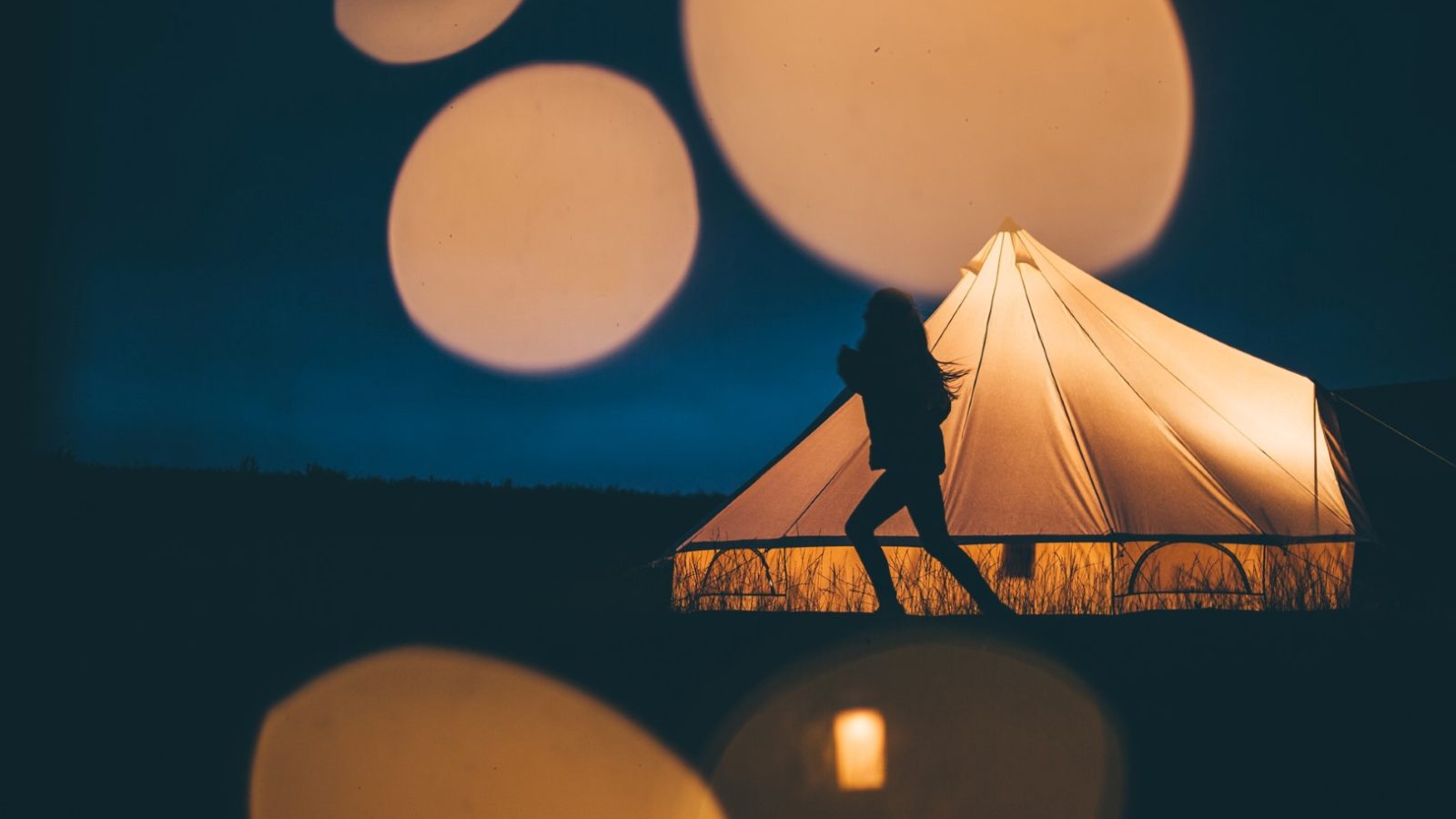 A silhouetted person walking near an illuminated tent at dusk captures the serene beauty of Nantseren. The sky is a mesmerizing dark blue, and large, blurred lights in the foreground create a dreamy atmosphere.