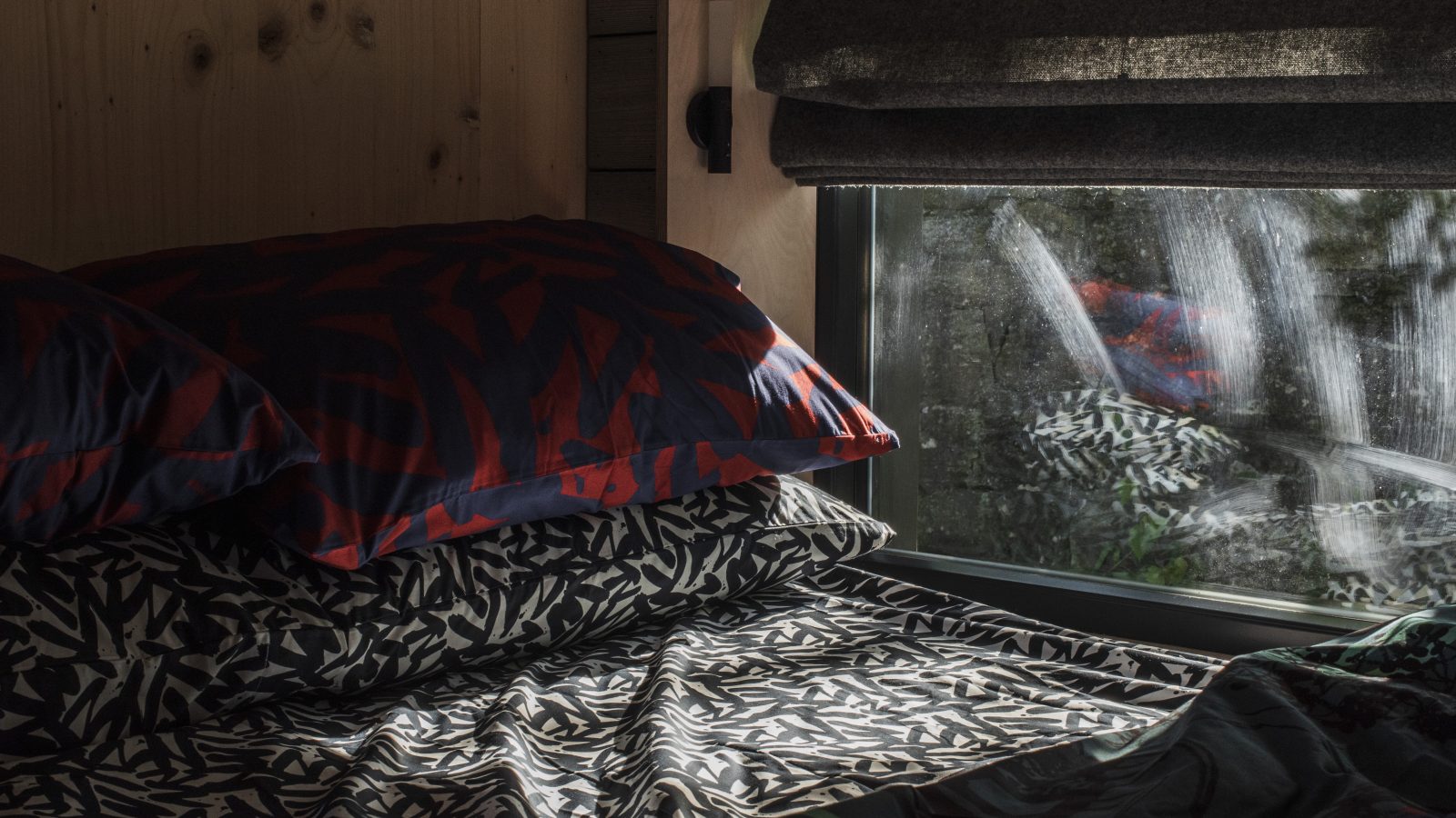 A cozy bed with patterned sheets and pillows sits beside a window, offering a serene view of greenery at Cambo Estate, perfect for an off-grid travel experience.