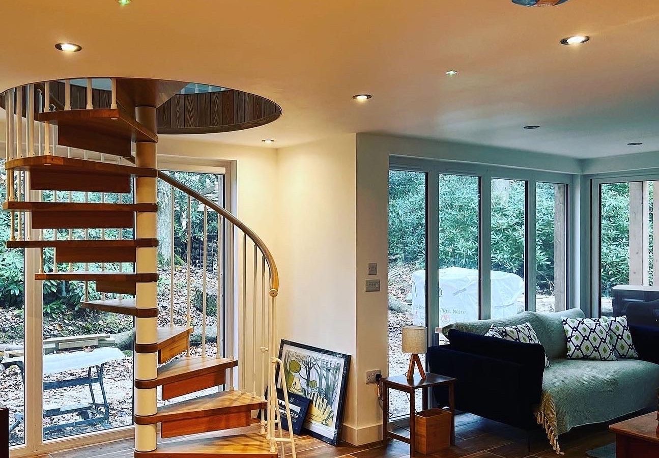 A cozy modern living space featuring a wooden spiral staircase, glass doors, and ample natural light. The room includes a sofa with patterned cushions, a small wooden table with a Coppertop Wood lamp, and framed artwork resting against the wall. A patio area with chairs is visible outside The Cabin.