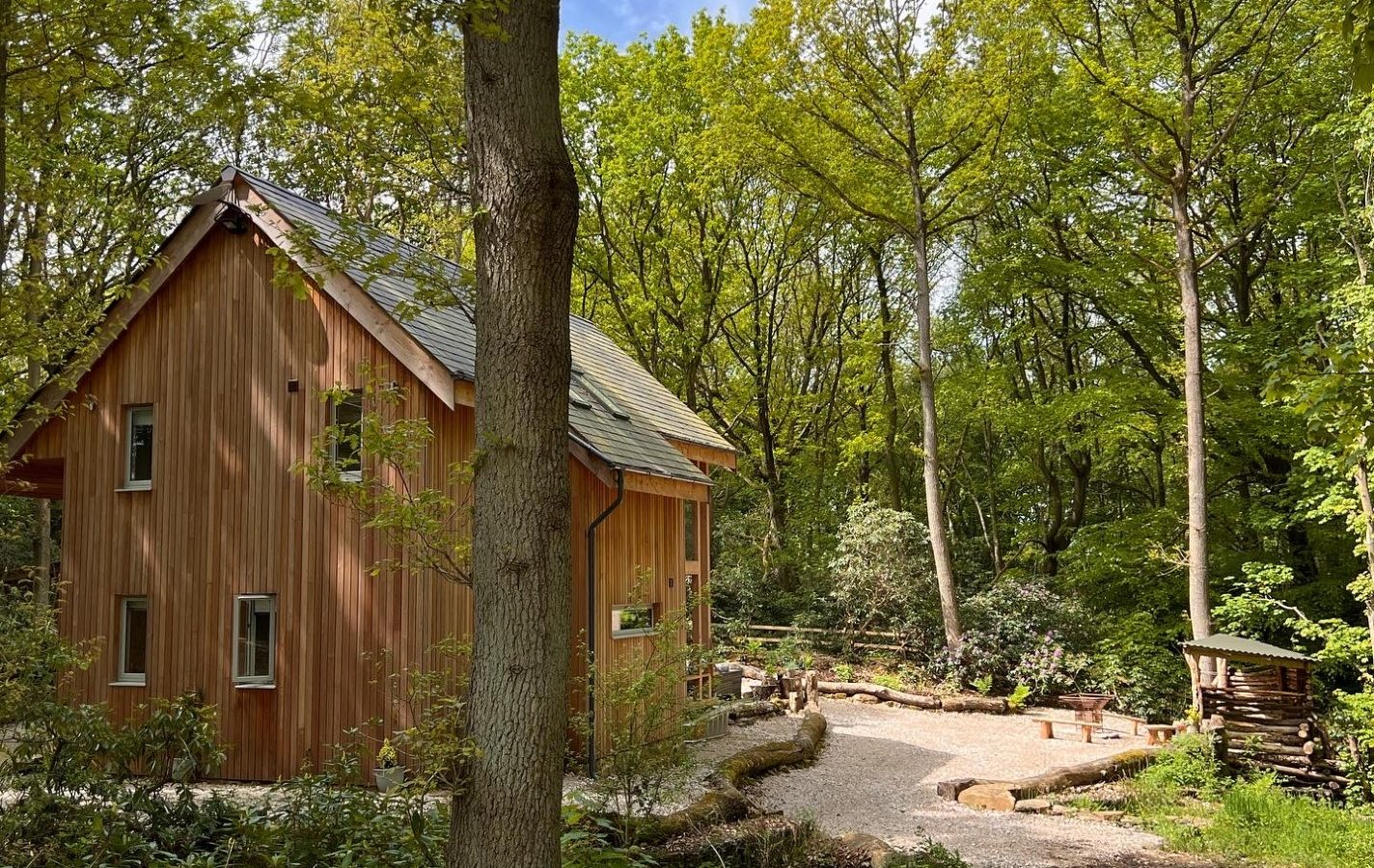 A Coppertop Wood cabin with a slate roof is nestled in a lush forest. It features a small, gravel-covered area with rustic benches surrounded by tall trees and greenery. The sunlight filters through the leaves, creating a serene atmosphere.