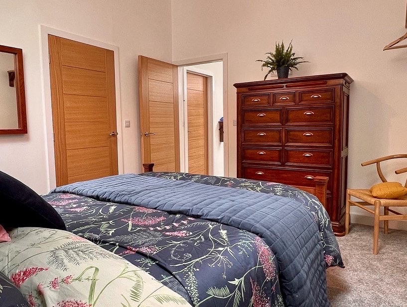 A cozy cabin bedroom featuring a bed with floral patterned bedding and a blue quilt. There are two wooden doors, a large Coppertop Wood dresser, a mirror, and a wooden chair. A potted plant is placed on top of the dresser. Neutral carpet covers the floor.