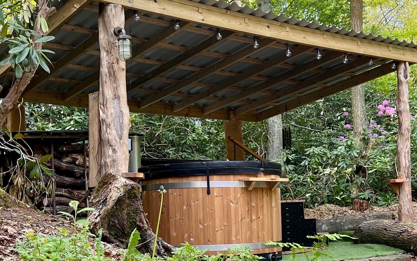 A wooden hot tub sits under a rustic Coppertop Wood canopy in a lush forest setting. Supported by natural tree trunks and adorned with hanging lights, it exudes a cozy cabin charm. Surrounding the area are green plants, bushes, and trees, creating a serene and natural atmosphere.