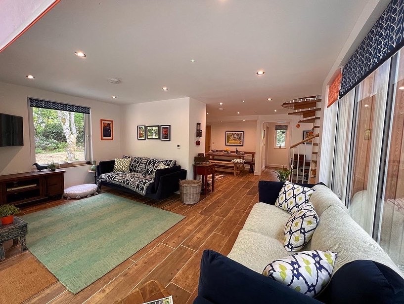 A cozy living room featuring two sofas with patterned cushions, a green rug, a TV on a wooden stand made of rich wood, and several framed pictures on the walls. A spiral staircase leads to the upper floor, and large windows allow natural light to fill the space, evoking a charming cabin feel.