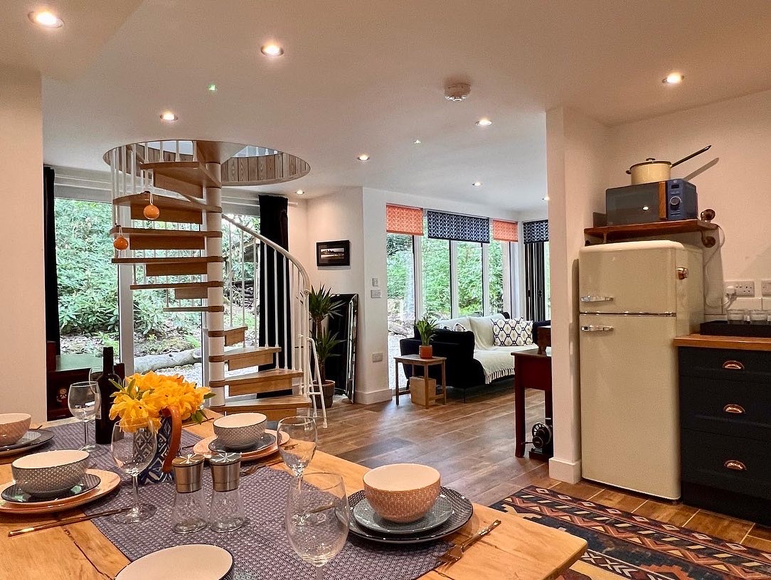 A modern, open-plan living space in The Cabin featuring a dining table set with plates, glasses, and yellow flowers in a vase. A spiral staircase leads to an upper floor. The room has large windows with views of greenery, a Coppertop Wood refrigerator, and cozy seating areas.