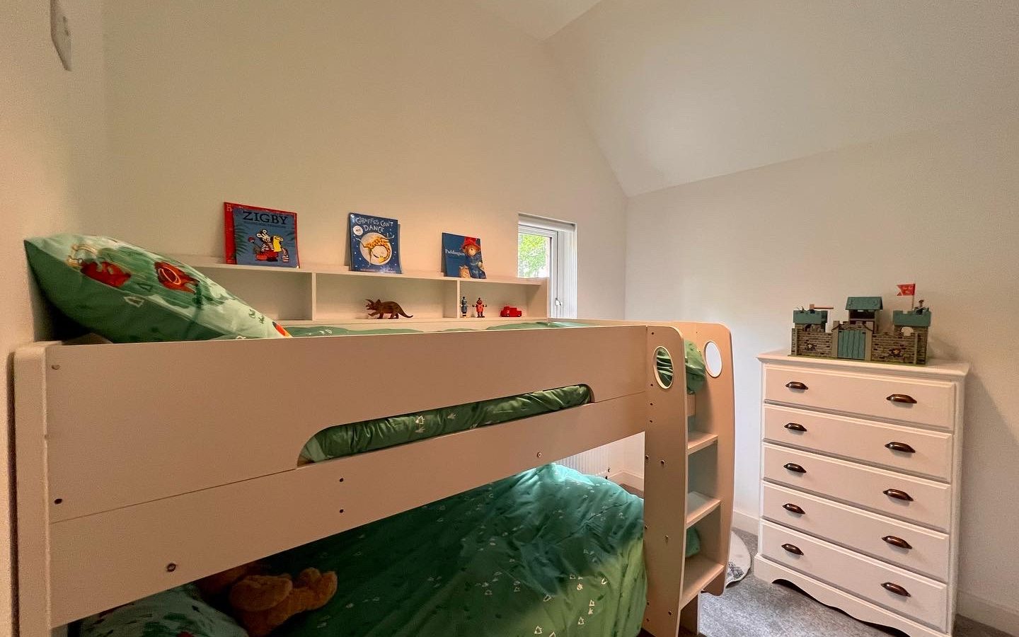 A children's bedroom with a white bunk bed, decorated with green bedding featuring dinosaur prints. The upper bunk has books and toys, while the lower bunk is partially visible. A white dresser with more toys and books is beside the bunk bed, all set in a cozy cabin-style room painted white.