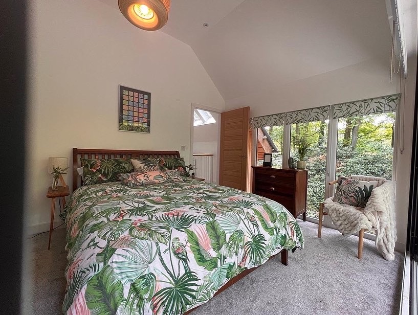 A bedroom with a high ceiling features a bed covered in leafy-patterned bedding. To the right is a Coppertop Wood dresser with a plant and a chair draped with a fluffy white blanket. Large windows reveal a green, leafy outdoor scene, and colorful artwork hangs on the wall, enhancing the cabin-like charm.