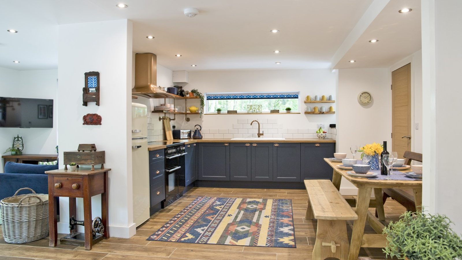 The bright open kitchen and dining area exudes a cabin-like charm with wooden floors. Featuring navy cabinets, a patterned rug, and a coppertop wood dining table with benches, the decor includes plants, wall art, and a window with a scenic view. Modern stainless steel appliances complete the look.