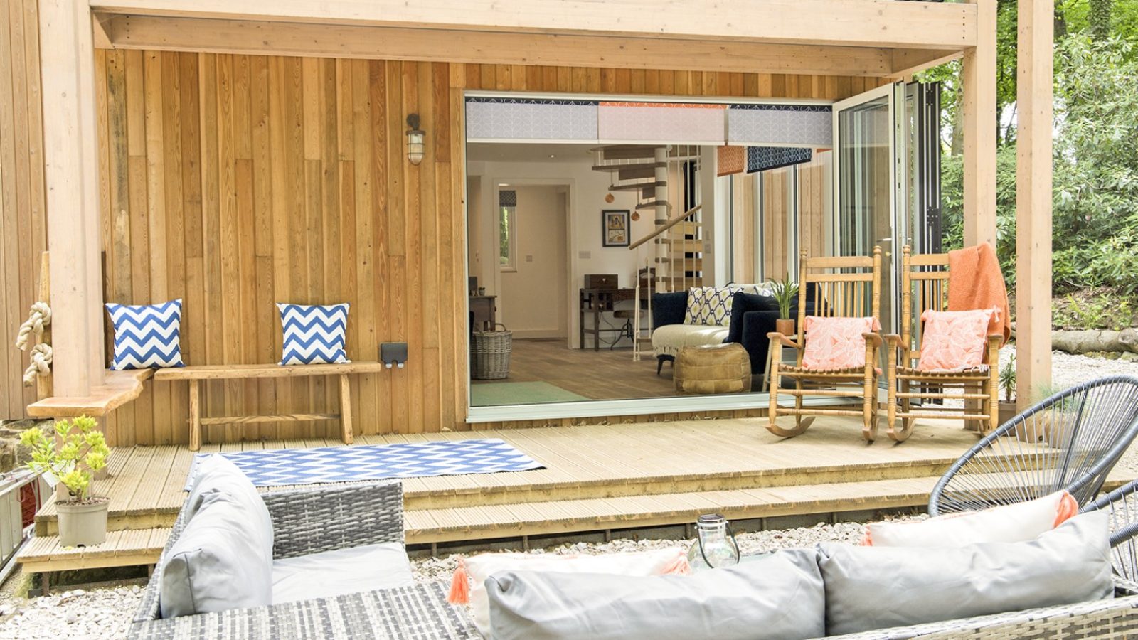 A modern outdoor deck with a Coppertop Wood facade, featuring rocking chairs and patterned cushions, overlooks a patio with wicker seating. Glass doors open to an interior with a visible staircase and cozy seating. The scene is surrounded by lush greenery, reminiscent of a serene cabin retreat.