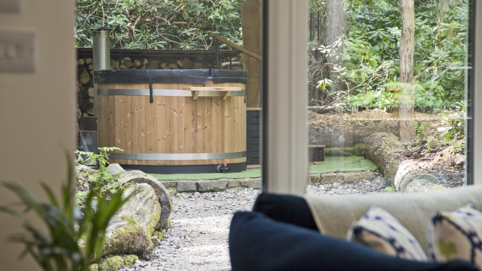 A cozy outdoor wooden hot tub is seen through a large window from inside The Cabin. The hot tub, nestled among greenery, logs, and stone, creates a rustic natural setting. Inside The Cabin, there is a blue sofa with patterned pillows.