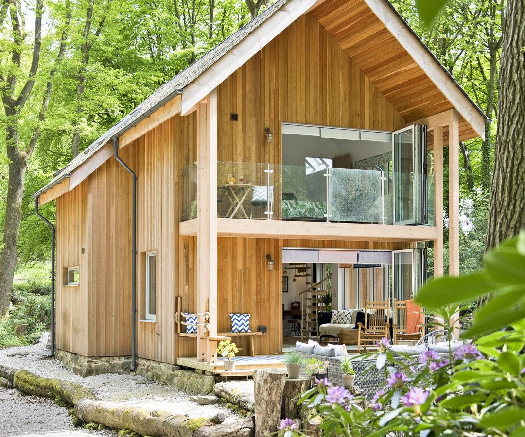 A modern two-story wooden cabin in a lush forest, featuring large glass windows and an open balcony. The surrounding area is filled with green trees and blooming purple flowers in the foreground, all harmonizing with the rustic charm of Coppertop Wood.