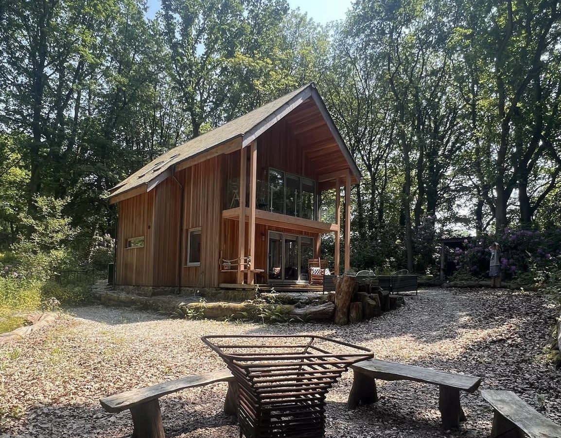 A modern two-story wooden cabin nestled in a forested area, known as Coppertop, features large glass windows and a covered porch. In the foreground, there's a circular outdoor seating area with wooden benches surrounding a metal fire pit. Sunlight filters through the trees.