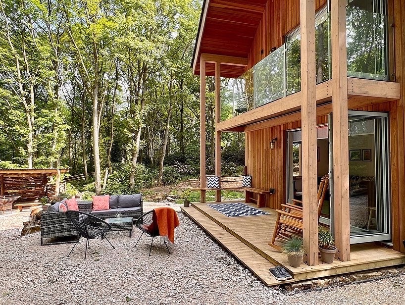 A modern Coppertop Wood cabin surrounded by trees features a spacious outdoor seating area with wicker furniture and colorful cushions. The cabin has a large porch with a wooden swing and floor-to-ceiling windows that offer a view of the forest. A shed is visible in the background.