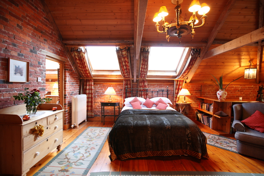 A cozy attic bedroom with warm wooden interiors and brick walls at Pennard Farm Cottages. The room features a double bed with red and pink pillows, two skylights, a wooden dresser adorned with flowers, a comfortable armchair, and a bookshelf. Soft lighting creates a warm and inviting atmosphere.