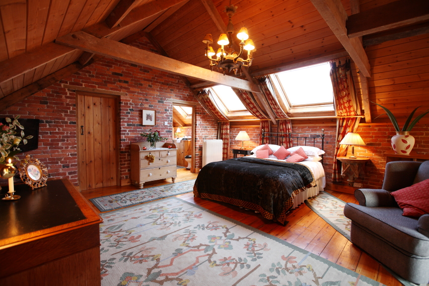 A cosy attic bedroom at Pennard Hill Farm Cottages featuring wooden beams, a brick wall, and skylight windows. The room has a large bed with pillows, a rug, a dresser, and a seating area with a sofa and lamps, creating a warm and inviting atmosphere. A chandelier hangs from the ceiling.