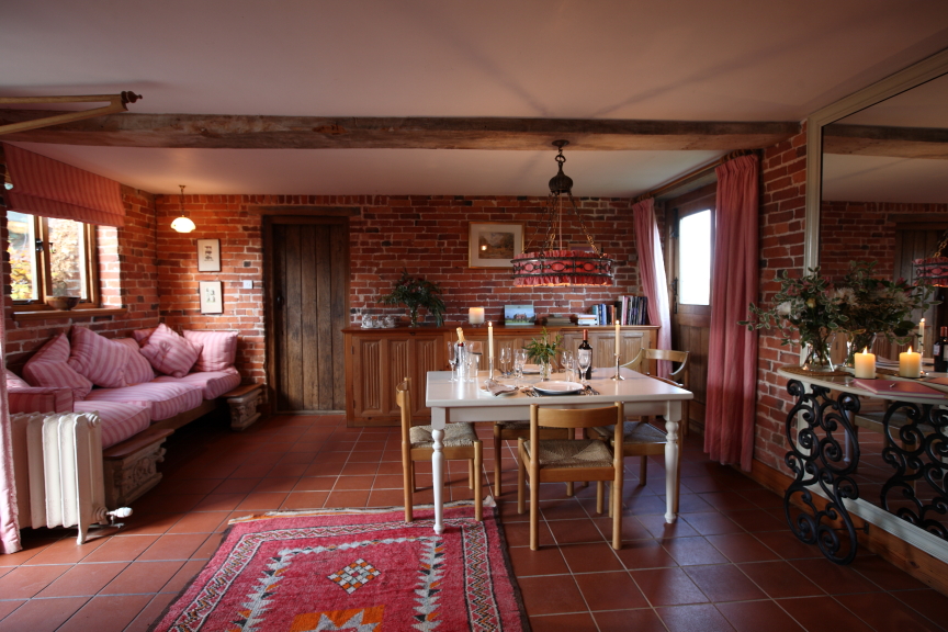 A cozy living and dining area with brick walls at Pennard Farm Cottages. The room has a wooden dining table with chairs, a cushioned bench, a sideboard with books and decor, a mirror, and plants. A colorful rug and candles add warmth and charm to the space. Large windows bring in natural light.