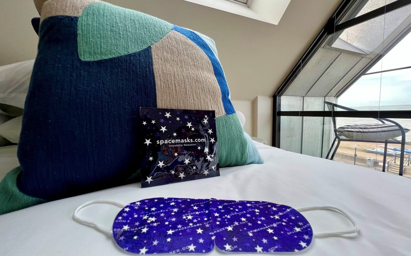 A purple sleep mask with a star pattern is placed on a white bed. Behind it, there is a matching purple package labeled 