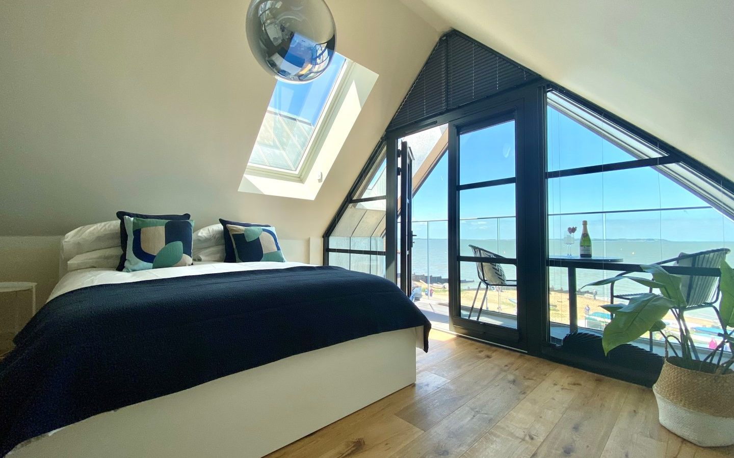 A modern attic bedroom with large windows offering an ocean view. The room features a double bed with geometric-patterned pillows, a small table and two chairs by the window, a potted plant on the wooden floor, and a hanging spherical light fixture to complete the look.