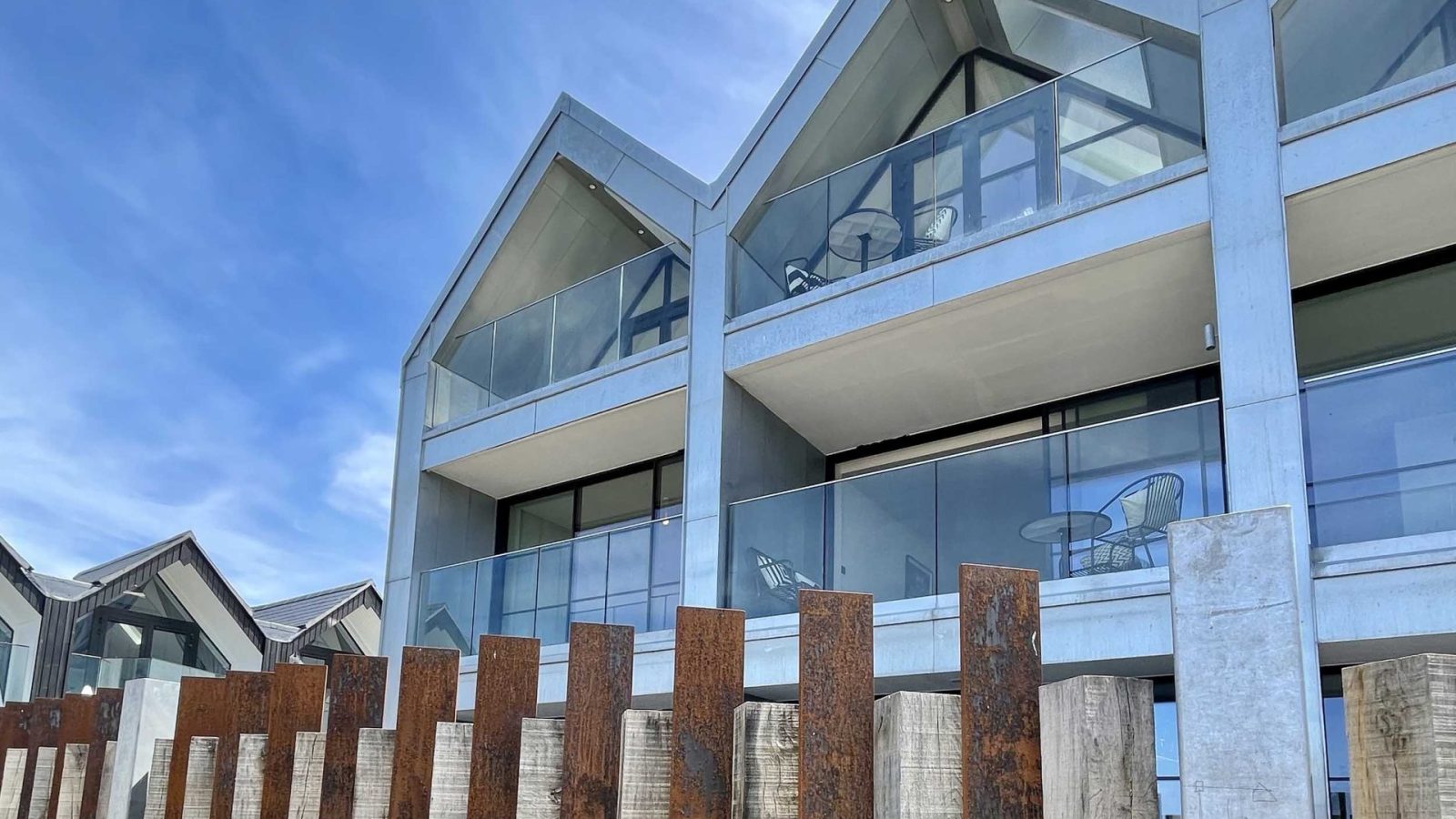 The Look Out is a modern building with a facade of large glass windows and balconies. The structure features pointed rooftops. In the foreground, there's a unique wooden fence with vertical panels of varying heights and some landscaped greenery. The sky is clear and blue.