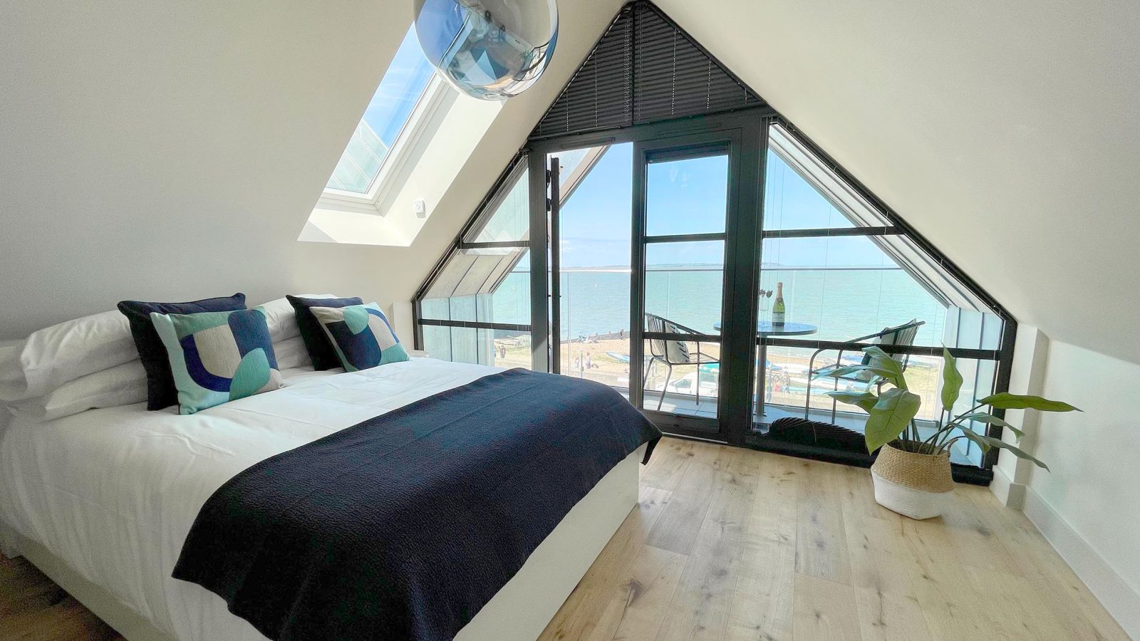 A cozy bedroom, known as The Look Out, features a large bed adorned with geometric cushions and a dark throw. Slanted ceilings and a floor-to-ceiling window open to a balcony with ocean views. A potted plant sits on the wooden floor near the window.