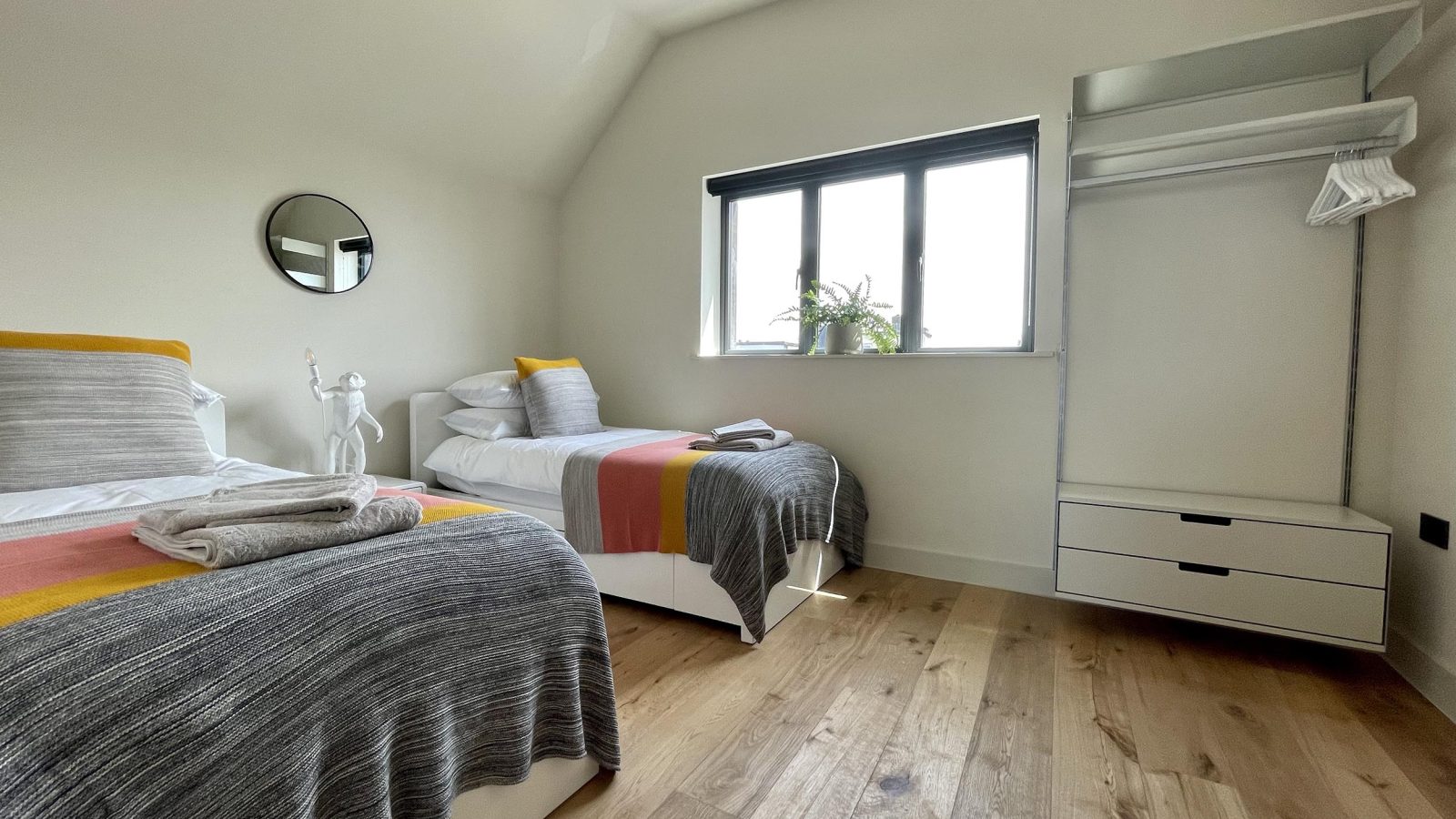 A modern bedroom with two single beds adorned with colorful striped blankets and neatly folded towels. The room, known as The Look Out, features a large window with a plant on the windowsill, hardwood floors, a round mirror on the wall, and an open wardrobe with drawers and hangers.