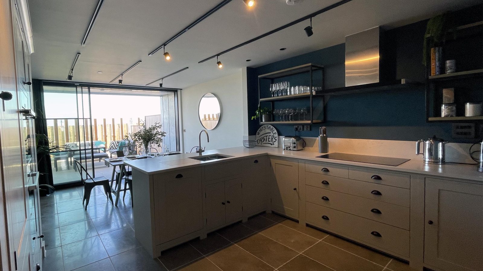 Modern kitchen with dark tile flooring, light gray cabinets, and a central island. Stainless steel appliances, a round mirror on the wall, and large windows offering a view of The Look Out's outdoor seating area are visible in the background. Shelves hold dishes and glassware.