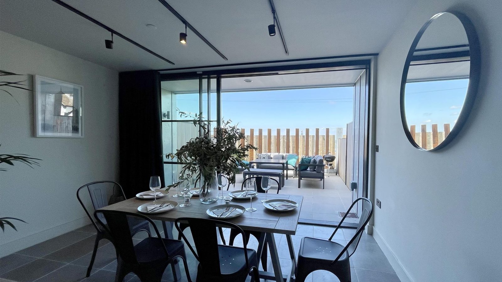 Modern dining area with a table set for four featuring white plates, black chairs, and a central floral arrangement. Large glass doors behind open to an observation point on the terrace with outdoor seating and a tall wooden fence. A circular mirror and framed picture adorn the walls.