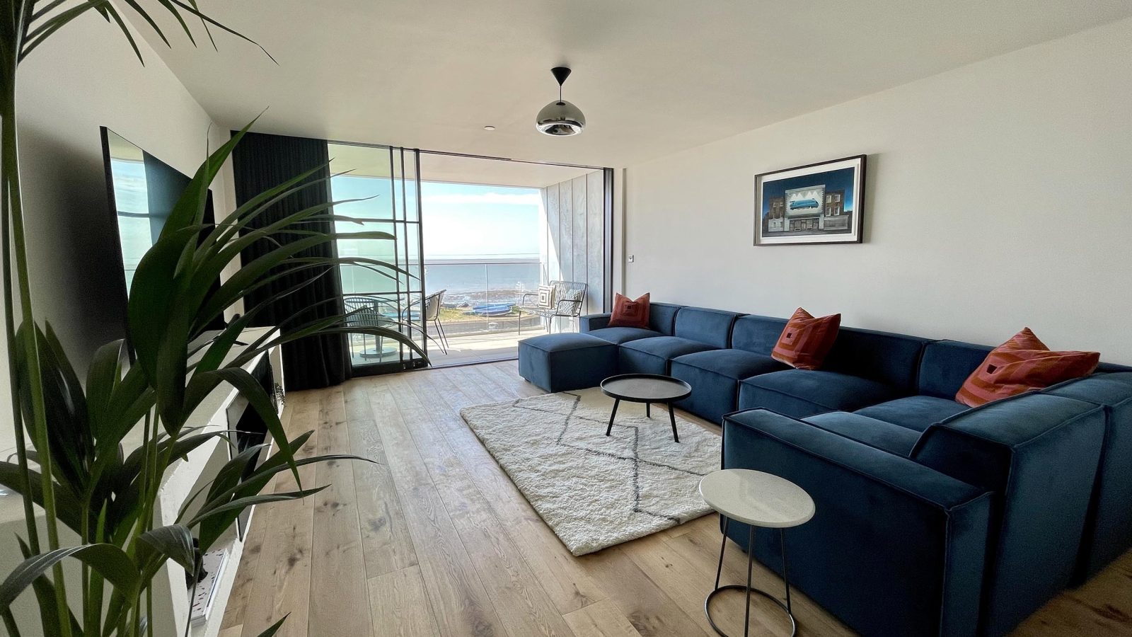 The Look Out features a modern living room with a large blue sectional sofa adorned with red throw pillows, a white area rug, and two small round tables. The bright room boasts wooden floors and a potted plant, complemented by a glass wall that opens to a balcony overlooking an ocean view.