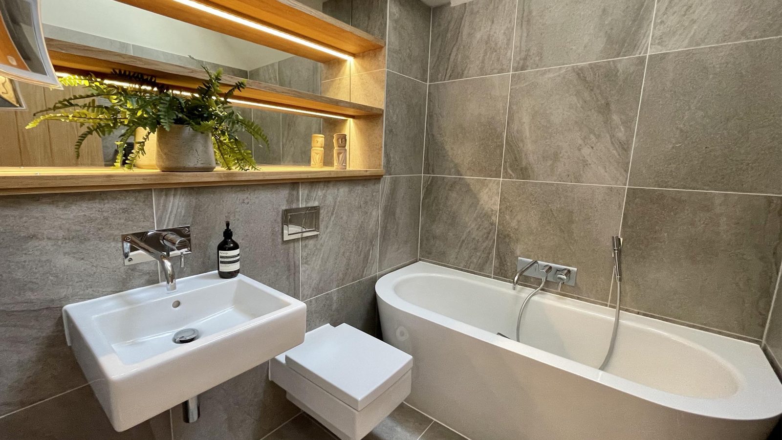 A modern bathroom featuring a wall-mounted sink, a floating toilet, and a freestanding bathtub with a wall-mounted faucet. Look out for the built-in shelf adorned with potted plants and toiletries. The room is decorated with large, stone-gray tiles and has sleek, contemporary fixtures.