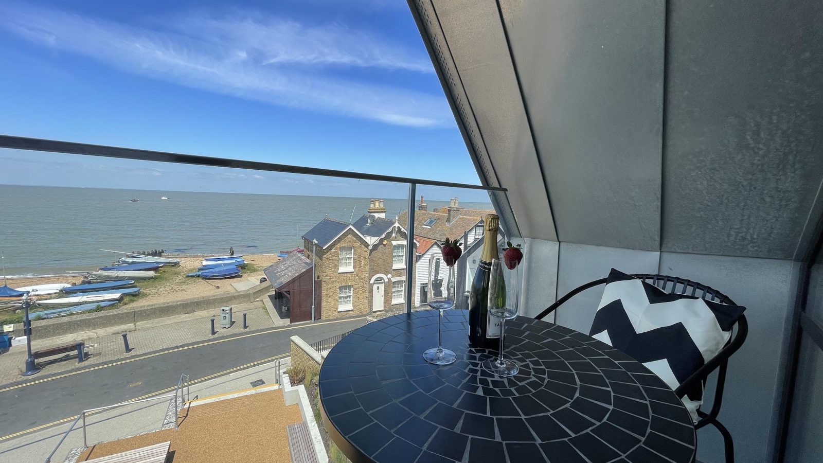 A balcony named The Look Out features a round black table and two chairs, offering a stunning seaside view. On the table are a bottle of wine, two wine glasses, and two roses. The scene includes a beach with boats, a road, and houses under a blue, partly cloudy sky.