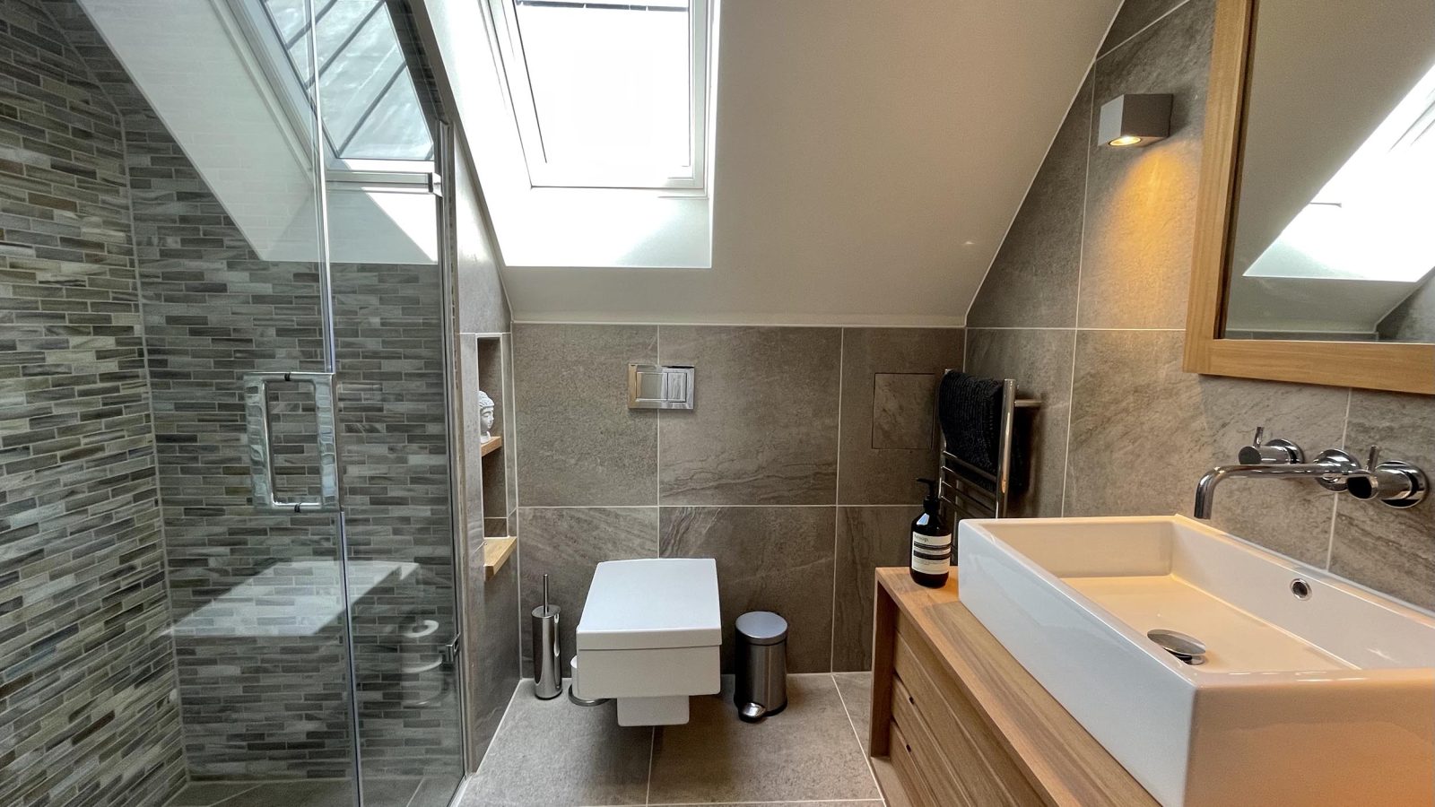 A modern bathroom in The Look Out features a sloped ceiling, glass-enclosed shower with a mosaic tile design, wall-mounted toilet, wooden vanity with a white sink and wall-mounted faucet, rectangular mirror, and skylight offering a scenic view that brings in natural light.